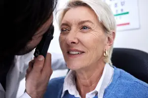A doctor checks an elderly woman's eyes with a handheld ophthalmoscope.