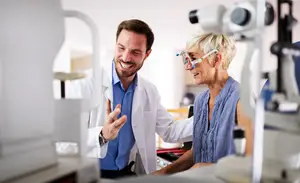 A smiling male doctor wearing a white coat is examining a female patient's eyes with a medical device while she wears glasses