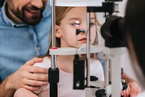 An image of a girl is sitting on a chair and looking into a device with a man behind her.