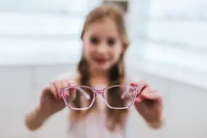 A girl wearing a pink shirt is holding up a pair of glasses with pink frames.
