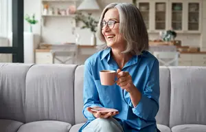 An elderly woman sitting on a couch, smiling and holding a cup of coffee.