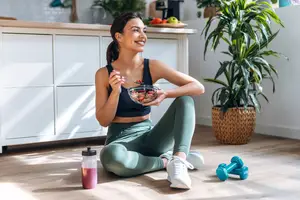A woman is sitting on the floor, smiling and holding a bowl of food in her hand.