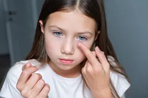 A girl with brown hair and blue eyes is putting in her contact lenses while looking at the camera with a serious face.