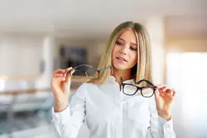 Blonde woman in white shirt holding up two pairs of glasses.