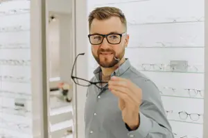 A man wearing glasses holding a pair of glasses in a store with shelves of glasses in the background.
