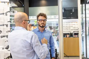 A man is trying on a pair of glasses while talking to a salesperson in a glasses shop.