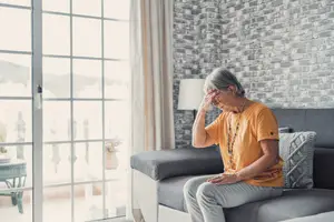 An elderly woman sitting on a couch with her hands on her head, looking sad.