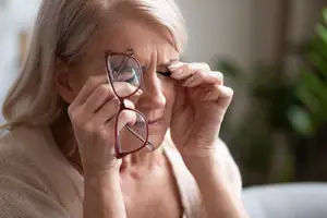 An older woman with blonde hair wearing glasses and a beige cardigan is looking at something with a serious expression and her hands are covering her eyes