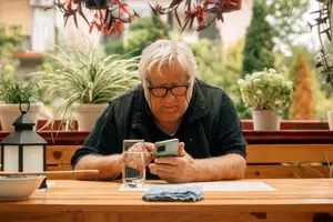 Old man wearing glasses and a black jacket is sitting at a wooden table outdoors, holding a cell phone and looking at the screen, with a glass of water and a lantern on the table, and potted plants and trees in the background.