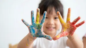 A young girl with her hands covered in paint is posing for a picture with a big smile on her face