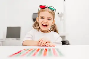 A young girl wearing 3D glasses is sitting at a table with colorful objects in front of her, smiling and looking at the camera.