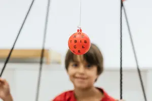 A child with short brown hair smiles while holding a red ball with letters on it that is hanging from a pulley system.