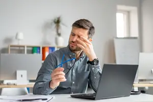 A man sitting at a desk with a laptop, rubbing his forehead, holding glasses in his hand, and looking at something on his laptop.