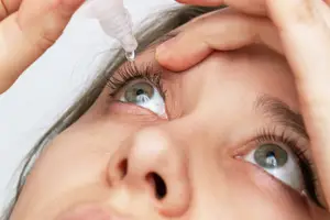 A woman applies eye drops to her eye, holding the dropper bottle with one hand and her eyelid open with the other.