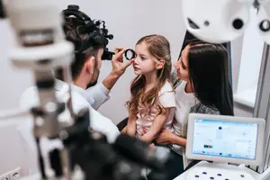 A young girl is having her eyes checked by a doctor while a woman looks on