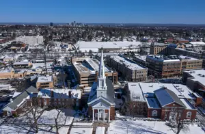 Atop a hill, an aerial view of a city covered in snow. Many buildings are visible, including a church, a parking garage, and a large building with a white roof. The city is surrounded by a large, white, snow-covered field. In the distance, mountains and trees are visible.