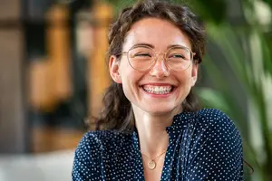 A smiling woman with brown curly hair wearing glasses and a polka dot blue blouse is looking at the camera with a couch and a potted plant in the background.