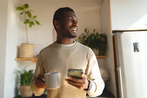 A smiling man holding a cup and a cell phone in a kitchen