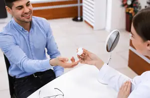 A man is receiving eyedrops from a woman in a white lab coat while sitting in a chair.