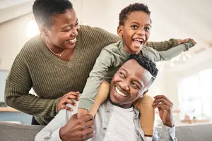 A woman and a man are smiling while playing with their son on a couch, and they are looking at the camera.