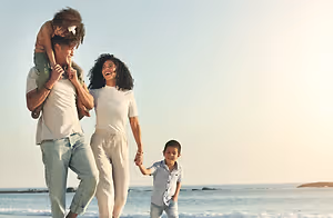 A happy family of three, with a woman holding hands with her two children on a beach.