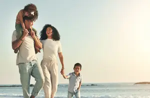 A happy family of three, with a woman holding hands with her two children on a beach.