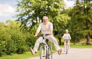 Two elderly people riding bicycles in a park, one of them smiling and jumping in the air