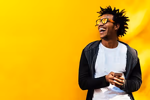 Man standing in front of a yellow wall, holding a phone, wearing sunglasses, jacket, and white shirt, with a smile on his face, looking at something.