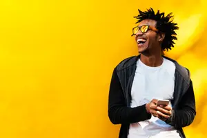 Man standing in front of a yellow wall, holding a phone, wearing sunglasses, jacket, and white shirt, with a smile on his face, looking at something.