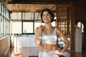 A woman wearing a white sports bra and white leggings is smiling and posing for a photo in a room with wooden floors and glass windows.