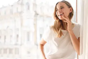 A woman standing near a window smiling with her hand on her chin