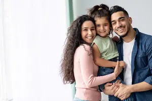 A smiling family of three stands together in a room, the woman holding a young girl, and the man holding the girl's hand while the girl looks on.
