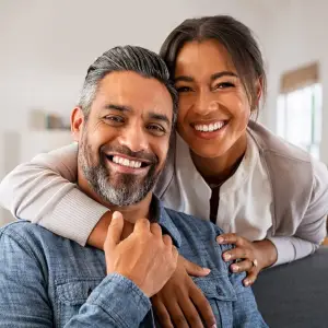 A smiling couple posing for a photo inside a room with a white wall and a window with blinds.