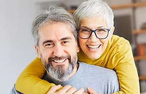An elderly woman with glasses hugging a man with a beard and gray hair in a room