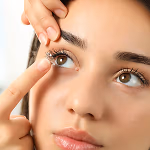 A woman is touching her eye with a finger and a lens on her finger. She has long eyelashes and is wearing makeup.