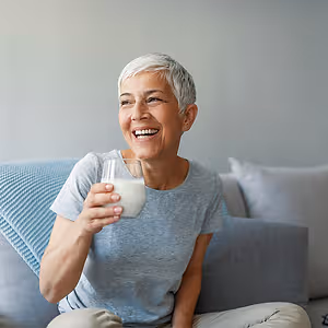 An older woman is sitting on a couch, smiling and holding a glass of milk in her right hand.