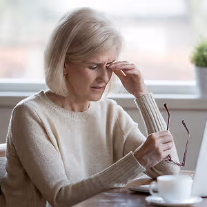 A woman in a beige sweater sitting at a table with a laptop and a cup of coffee looks stressed.