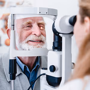 An elderly man is looking into an eye examination machine while a woman looks on in the background