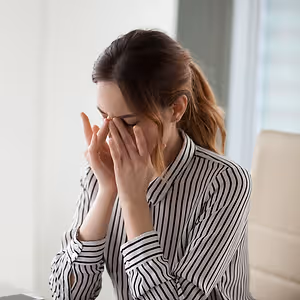 A woman in a striped shirt is sitting in an office chair, covering her face with her hands, and appears to be in distress.