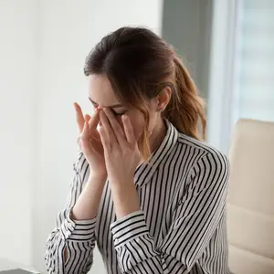 A woman in a striped shirt is sitting in an office chair, covering her face with her hands, and appears to be in distress.