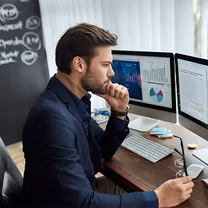 A man is sitting in front of a desk with a monitor, keyboard, mouse, and other items. He is looking at something while holding his chin with his right hand and is wearing a suit, a watch, and a ring.