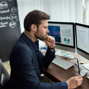 A man is sitting in front of a desk with a monitor, keyboard, mouse, and other items. He is looking at something while holding his chin with his right hand and is wearing a suit, a watch, and a ring.