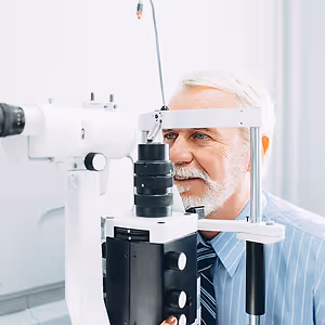 An older man is smiling while examining his eyes with an ophthalmoscope in a medical room.