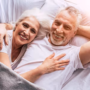 An elderly couple smiles and poses for a photo in their bedroom