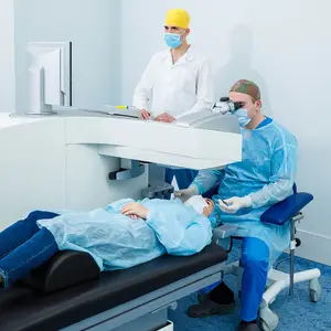 Three people in medical attire are in a hospital room, with one person lying on a bed and the other two standing and looking through a microscope.