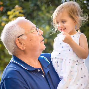 A happy old man is holding a young girl in his arms and looking at her in a garden area with trees and plants in the background.