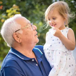A happy old man is holding a young girl in his arms and looking at her in a garden area with trees and plants in the background.