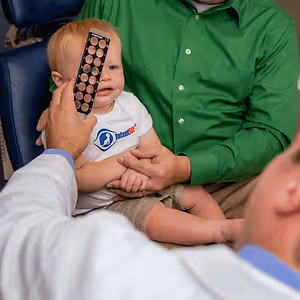 A young child is having their eyes examined by an adult male doctor while being held by a man in a green shirt.