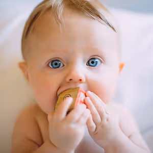 A baby with blue eyes is holding a wooden toy in its mouth while lying on a white surface.