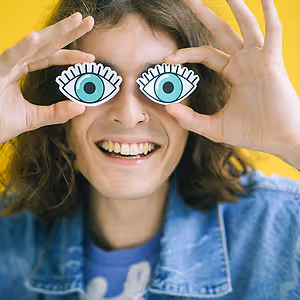 A smiling person wearing denim jacket and holding up glasses with green eyes and white circles
